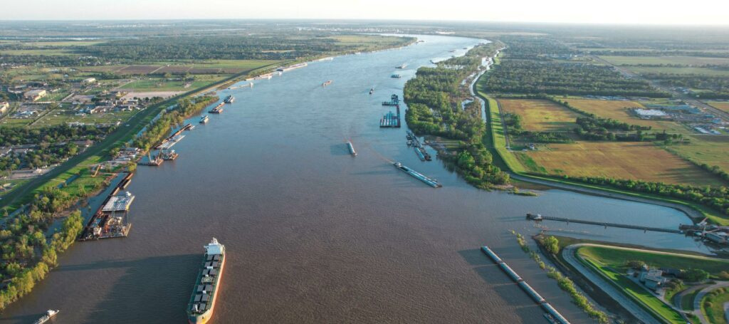 An aerial view shows a wide, winding river with several barges and ships, flanked by lush green land and industrial areas