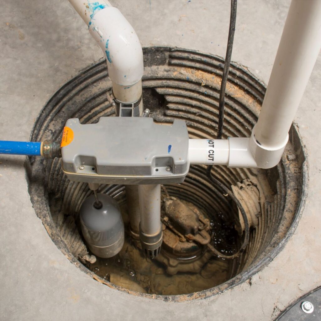 A sump pump system with white PVC pipes and a gray control unit is visible within a circular pit in a concrete floor, containing standing water