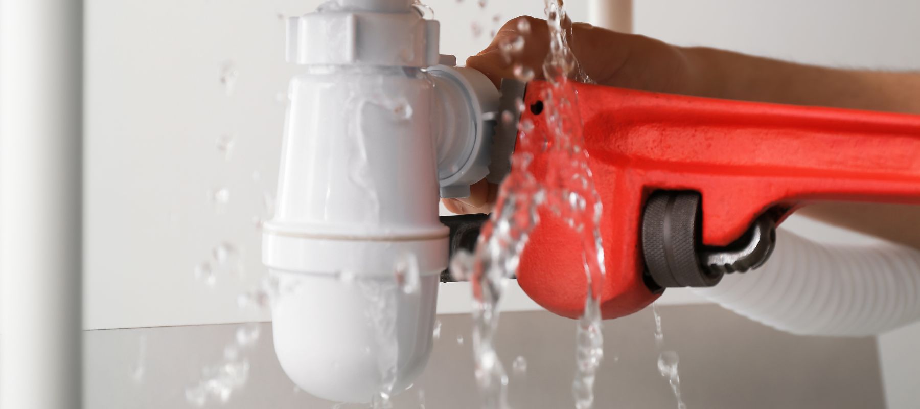 Water sprays from a white PVC pipe connection as a red wrench is used to tighten it, indicating a leak