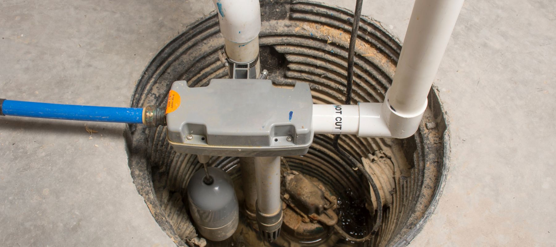 A sump pump system with white PVC pipes and a gray control unit is visible within a circular pit in a concrete floor, containing standing water