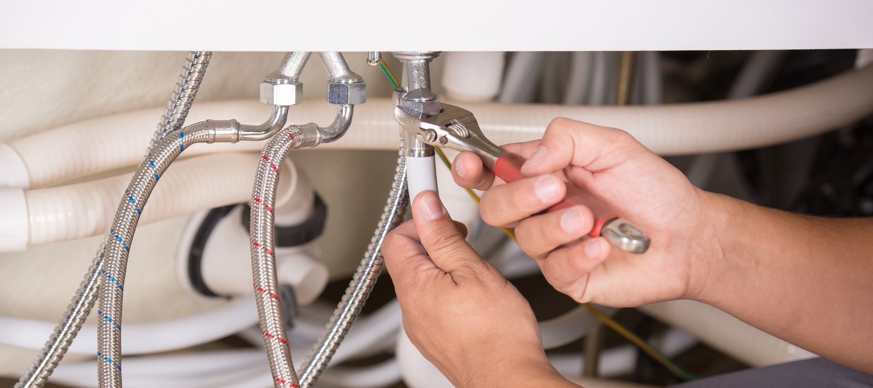 A plumber's hands use a wrench to tighten a pipe fitting among a cluster of braided and white pipes