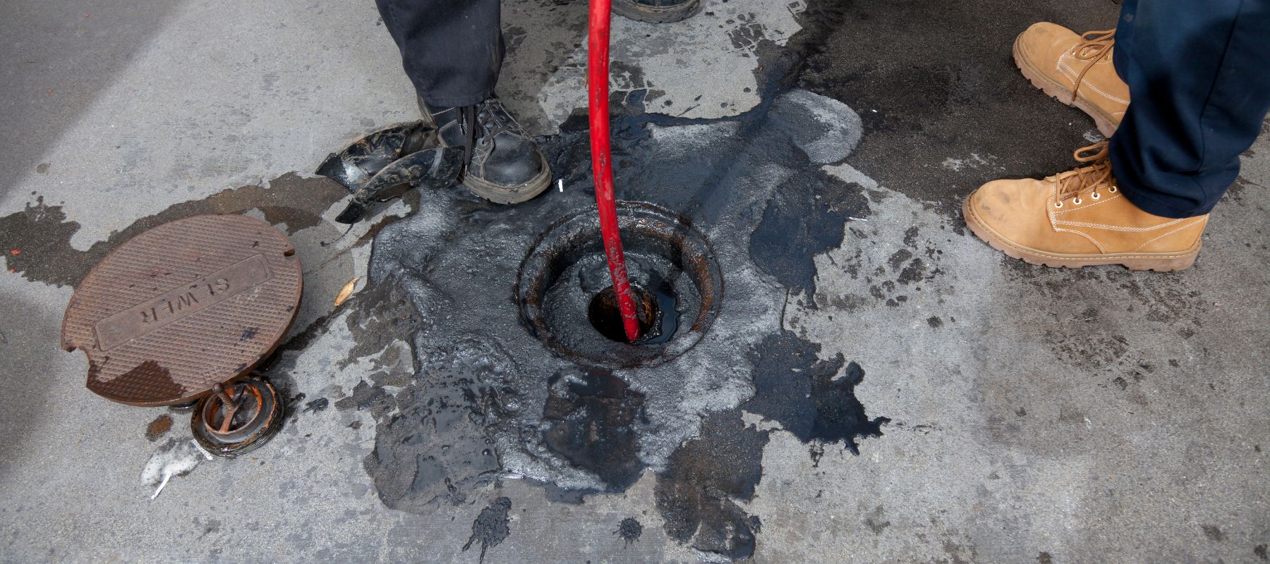 A bright red hose is inserted into a grimy, open drain on a concrete floor, surrounded by dark sludge
