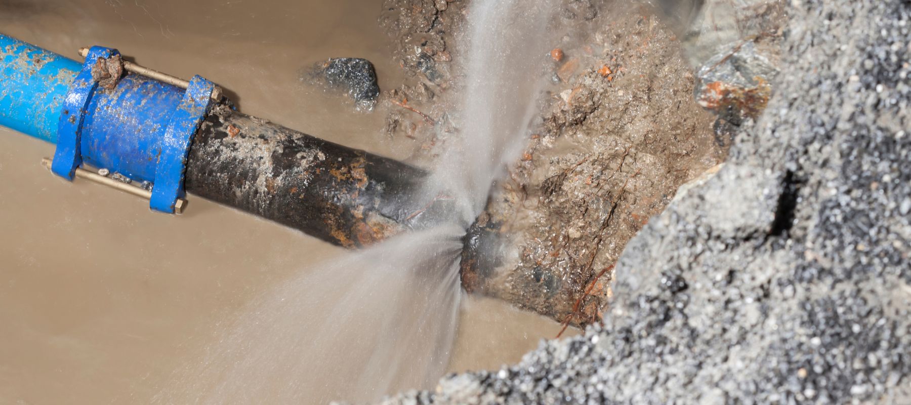 A large, dark pipe with a blue clamp violently leaks water from a crack into a muddy trench