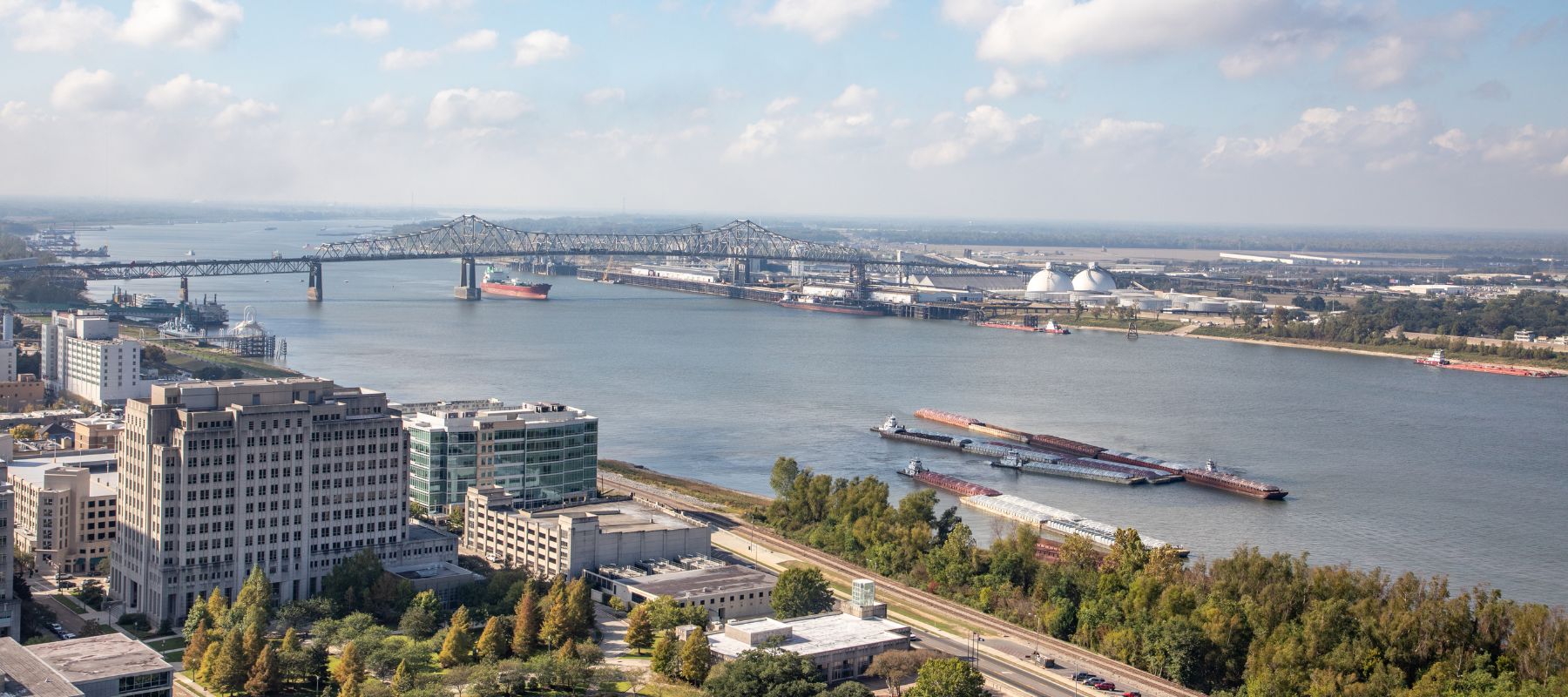 An aerial view captures a wide river with cargo ships and barges, a large bridge, and buildings lining the riverbanks under a partly cloudy sky