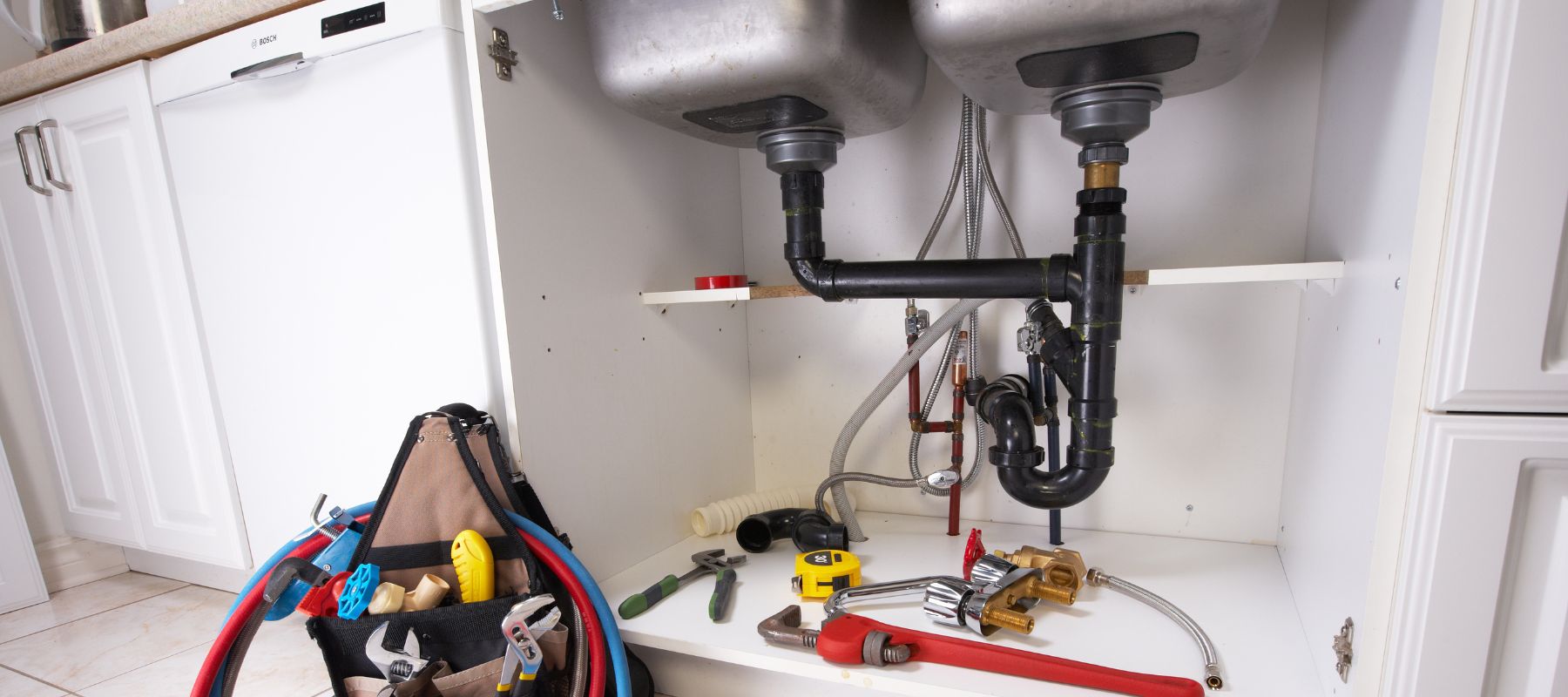 Plumbing tools are neatly arranged inside and around an open cabinet under a double kitchen sink, revealing the pipes and connections