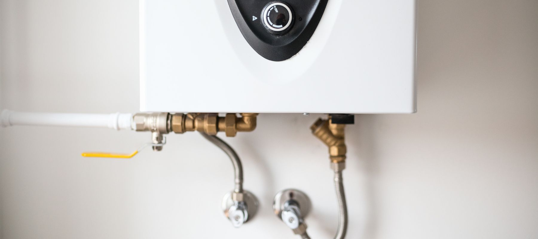 A white tankless water heater with a black control panel and a dial is mounted on a wall, connected to brass and chrome pipes