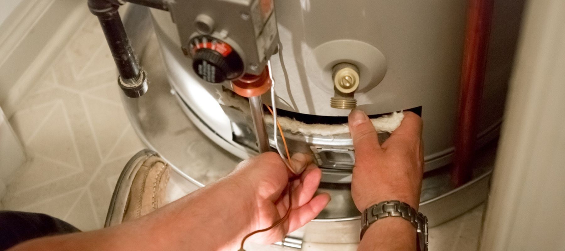 A person's hands work on exposed wiring and insulation at the base of a water heater with a thermostat.