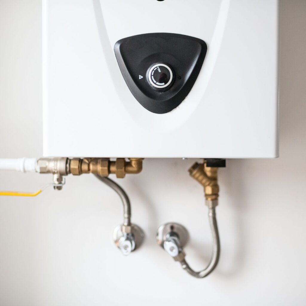 A white tankless water heater with a black control panel and a dial is mounted on a wall, connected to brass and chrome pipes