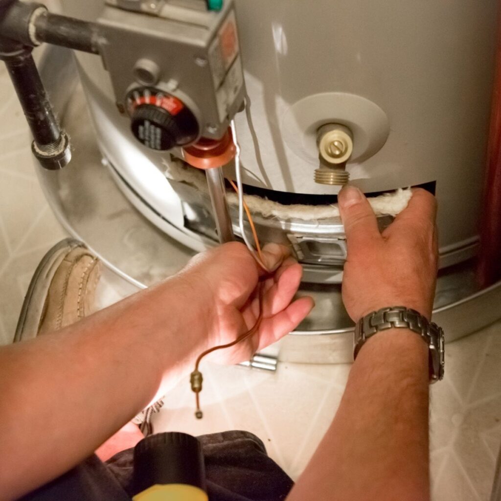 A person's hands work on exposed wiring and insulation at the base of a water heater with a thermostat