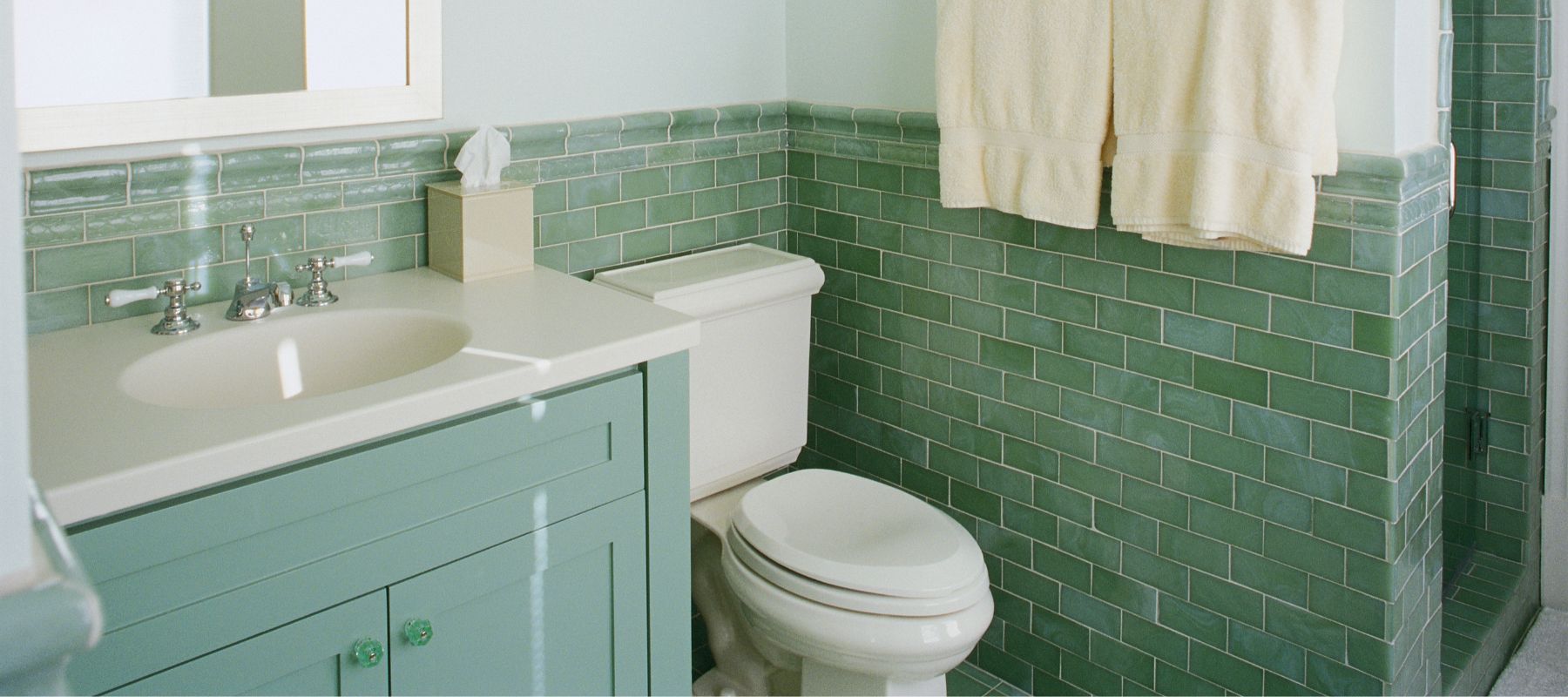 A vintage-style bathroom features a mint green vanity with a toilet and green subway tiles on the walls, with white towels hanging nearby